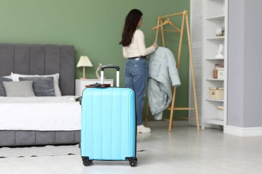Mavi bavul ve genç bir kadın evdeki elbise rafına ceket asıyor. Seyahat kavramı