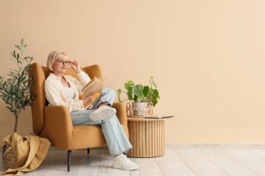 Bej duvarın yanındaki koltukta kitap okuyan orta yaşlı bir kadın.