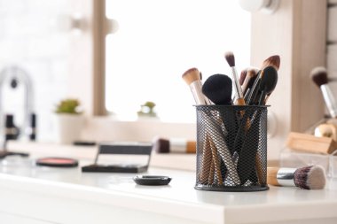 Makyaj fırçaları ve farklı dekoratif kozmetik ürünleri olan tutucu evdeki beyaz masada.