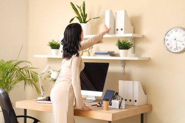 Çalışma yerinin yanındaki raftan modern bilgisayarla şık bir ofiste klasör alan genç bir kadın.