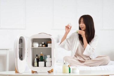Kozmetik ürünleri için mini buzdolabı ve yatakta güzel genç bir kadın.