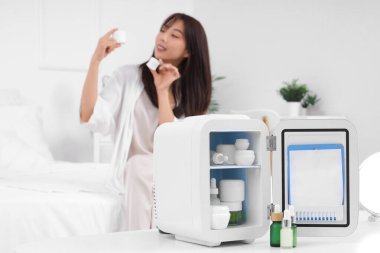 Masada küçük bir kozmetik buzdolabı ve evde yatakta güzel bir genç kadın.