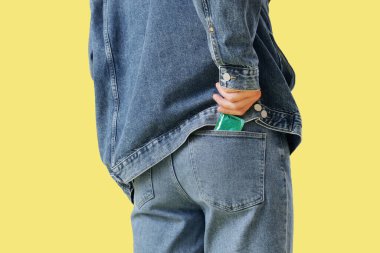 Genç adam sarı arka planda kot pantolon cebine prezervatif koyuyor.