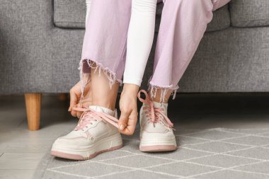 Spor ayakkabılı ve pembe pantolonlu genç bir kadın evde ayakkabı bağı bağlıyor.