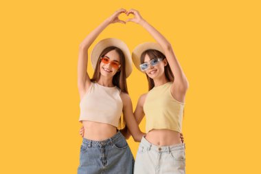 Güneş gözlüklü güzel genç kadınlar ellerini sarı arka planda kalp şeklinde yapıyorlar.