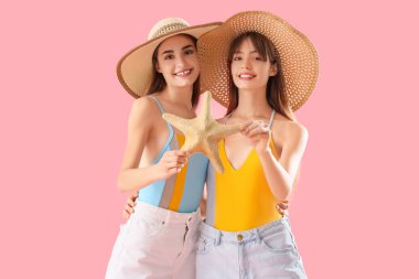 Hasır şapkalı güzel genç kadınlar pembe arka planda denizyıldızı tutuyorlar.