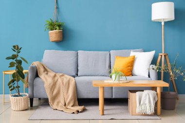 Mavi duvarın yanındaki yastıklı, ekoseli ve kahve masalı rahat bir kanepe.