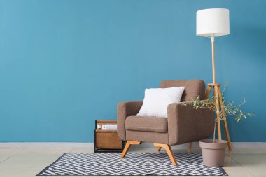 Konforlu koltuk, dekoratif zeytin ağacı, lamba ve dergi tutacağı mavi duvarın yanında.