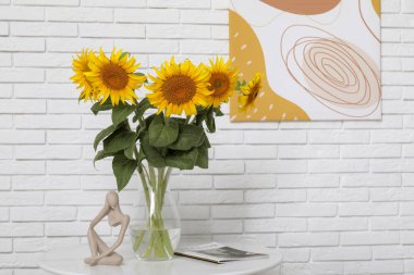 Oturma odasındaki beyaz tuğla duvarın yanındaki masada güzel ayçiçekleri olan heykel, dergi ve vazo.