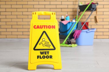 Temizlik malzemeleri, uyarı levhası ve tuğla duvarın yanındaki zemin paspası olan kapıcı vagonu.