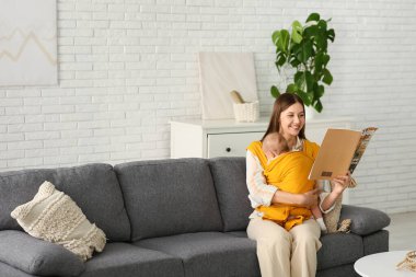 Kanepede oturan ve evde dergi okuyan küçük tatlı bir bebeği olan genç bir anne.