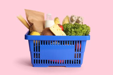 Pembe arka planda farklı taze ürünler ve peynirlerle dolu mavi alışveriş sepeti.