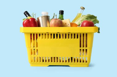 Farklı taze ürünlerle dolu sarı alışveriş sepeti ve mavi arka planda bir şişe meyve suyu.