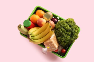 Farklı taze ürünlerle dolu yeşil alışveriş sepeti ve pembe arka planda muzlar. Üst görünüm