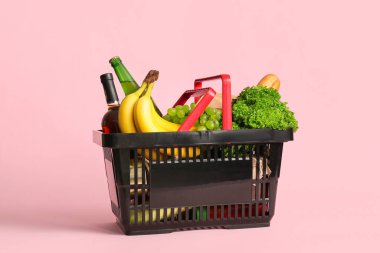 Farklı taze ürünlerle dolu siyah alışveriş sepeti ve pembe arka planda muzlar.