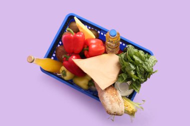 Farklı taze ürünlerle dolu mavi alışveriş sepeti ve leylak arka planında bir şişe yağ. Üst görünüm