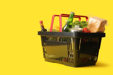 Farklı taze ürünlerle dolu siyah alışveriş sepeti ve sarı arka planda ekmek.
