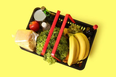 Farklı taze ürünlerle dolu siyah alışveriş sepeti ve sarı arka planda ekmek. Üst görünüm