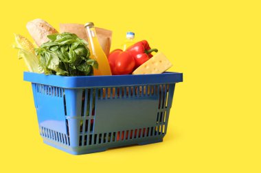 Farklı taze ürünlerle dolu mavi alışveriş sepeti ve sarı arka planda bir şişe meyve suyu.