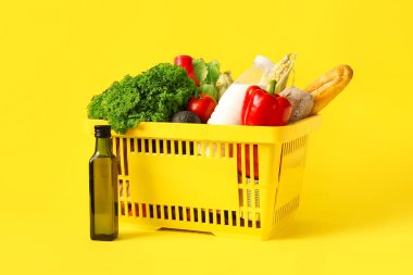 Bir şişe yağ, farklı taze ürünlerle dolu sarı alışveriş sepeti ve sarı arka planda dolma biber.