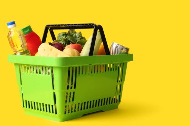 Farklı taze ürünlerle dolu yeşil alışveriş sepeti ve sarı arka planda bir şişe yağ.