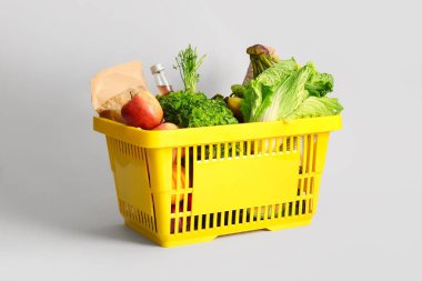 Beyaz arka planda farklı taze ürünler ve elmalarla dolu sarı alışveriş sepeti.