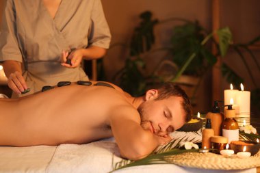 Genç adam masaj yaptırıyor. Koyu renk salonda sıcak spa taşlarıyla.