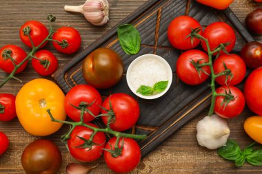 Farklı taze domatesler, fesleğen, sarımsak ve ahşap arka planda bir kase tuz.