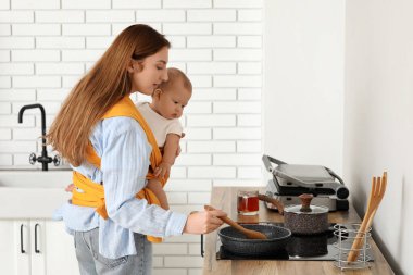 Genç anne mutfakta küçük bebekle yemek pişiriyor.