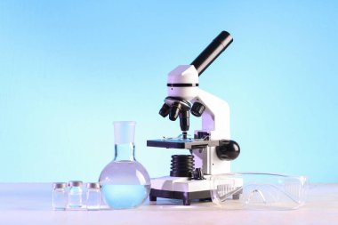 Modern mikroskop, ampuller, güvenlik gözlükleri ve beyaz masada sıvı bulunan laboratuvar şişesi.