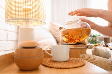 Kadın elleri çaydanlıktan fincana yeşil çay dökerken yatak odasındaki komodinin yanına lamba koyuyor. Yakın çekim.