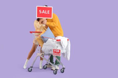 Mor arka planda alışveriş arabası ve satılık tabelaları olan genç bir çift.