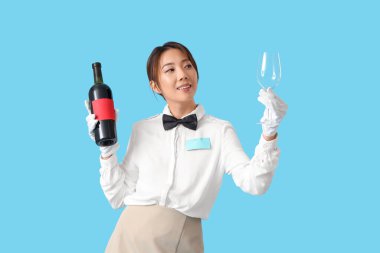 Mavi arka planda şarap ve cam şişesiyle gülümseyen genç Asyalı garson.