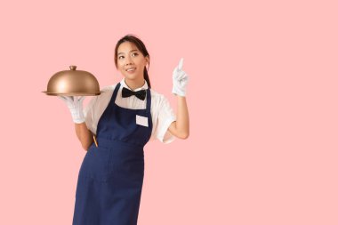 Yükseltilmiş işaret parmağı tepsisi ve pembe arka planı olan Asyalı genç bir garson.