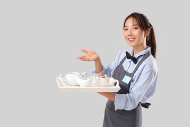 Genç Asyalı gülümseyen garson ve gri arka planda çay setiyle tepsi