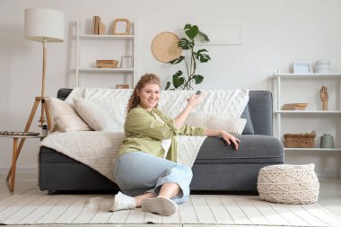 Klimalı, uzaktan kumandalı gülümseyen genç kadın evdeki kanepenin yanında oturuyor.