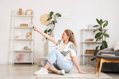 Klima uzaktan kumandalı güzel, gülümseyen genç bir kadın evde yerde oturuyor.