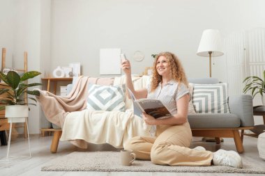 Klimalı, uzaktan kumandalı ve dergili güzel, gülümseyen genç bir kadın oturma odasında yerde oturuyor.