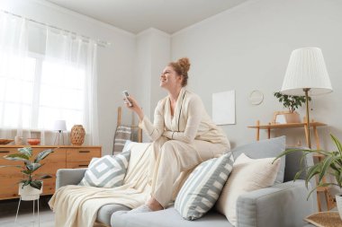 Klimalı, uzaktan kumandalı gülümseyen genç kadın evdeki kanepenin arkasında oturuyor.