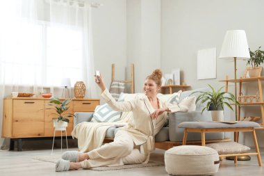 Uzaktan kumandalı ve kremli gülümseyen genç kadın oturma odasında.