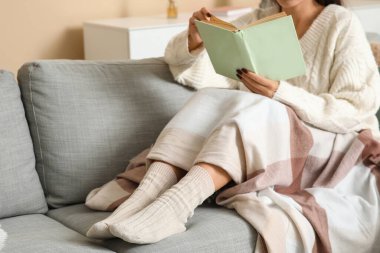 Sıcak çoraplı, battaniyeli ve kitaplı bir kadın evdeki kanepede oturuyor.