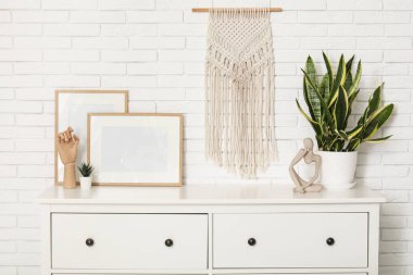 Beyaz tuğla duvarın yanındaki çekmecelerde fotoğraf çerçeveleri, biblo ve ev bitkisi.