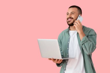 Bilgisayarlı genç adam pembe arka planda cep telefonuyla konuşuyor.