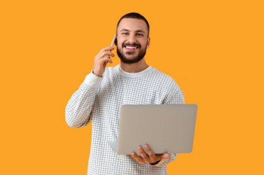 Laptoplu genç adam sarı arka planda cep telefonuyla konuşuyor.