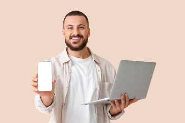 Bej arka planda cep telefonu ve dizüstü bilgisayarı olan genç bir adam.