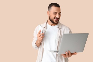 Bej arka planda cep telefonu ve dizüstü bilgisayarı olan genç bir adam.