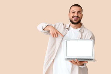 Genç adam bej arka planda dizüstü bilgisayara bakıyor.