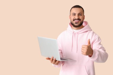 Dizüstü bilgisayarı olan gülümseyen genç adam bej arka planda baş parmağını gösteriyor.