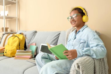 Kulaklıklı Afro-Amerikalı genç kız oturma odasında kanepede otururken kitapla sesli kitap dinliyor.
