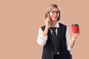 Başarılı bir iş kadını, bir fincan kahveyle bej arka planda cep telefonuyla konuşuyor.